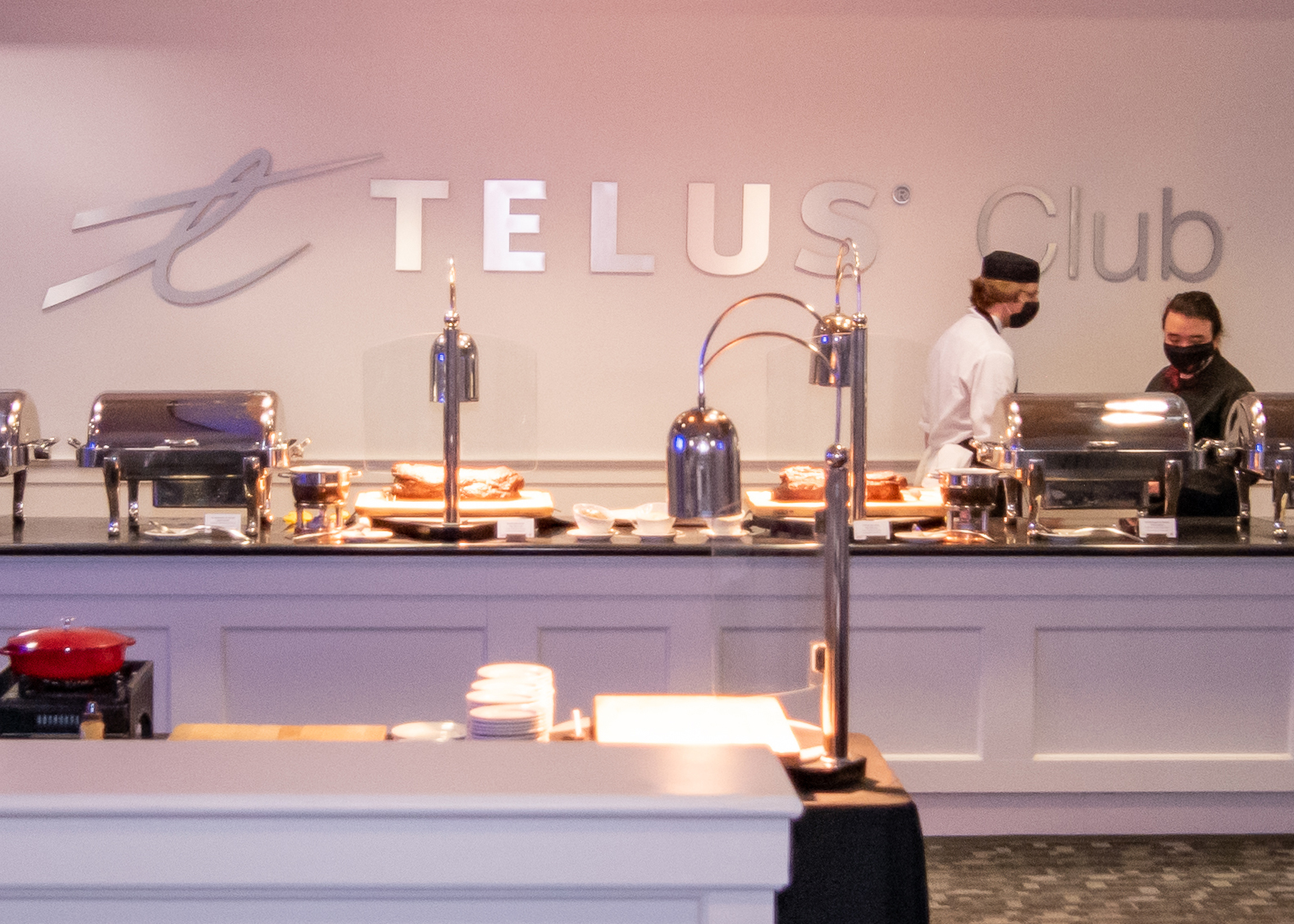 Inside the Telus Club, two chefs are standing behind the buffet table that is laid out with food, Telus Club logo is on the wall behind them