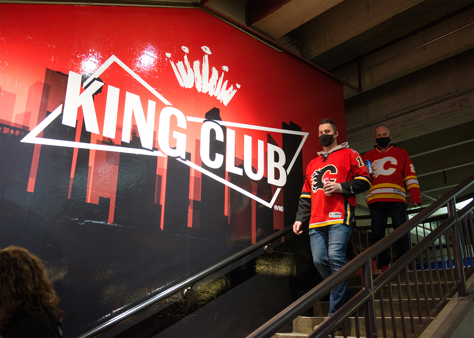 Two men wearing Calgary Flames jerseys walking down the stairs while a large King Club logo is on the wall next to them