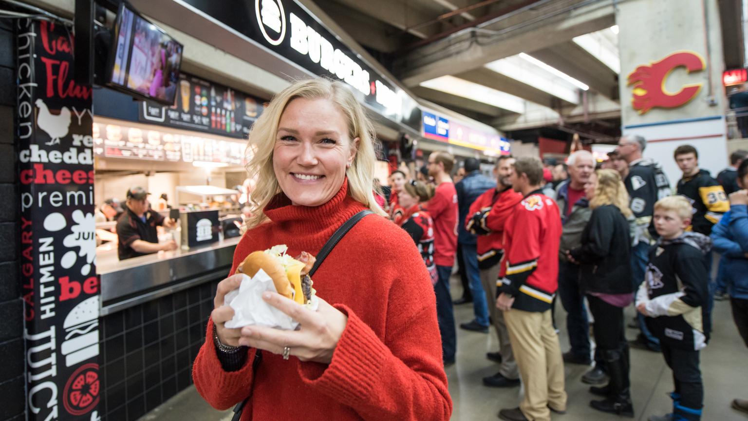 Woman holding a burger smiling, people lining up for food in the background